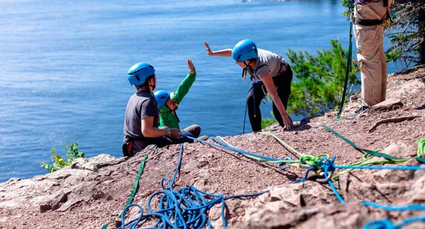 two people who are rock climbing high five just over the edge of a cliff. There is blue water below them.